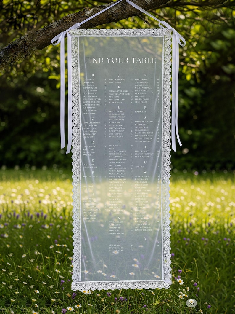 K&ouml;nnte beinhalten: Eine transparente Hochzeits-Sitzordnung mit Spitzenbord&uuml;re und dem Text "FIND YOUR TABLE". Die Tafel h&auml;ngt mit wei&szlig;en B&auml;ndern an einem Ast. Der Hintergrund ist eine verschwommene gr&uuml;ne Wiese mit Wildblumen.