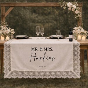 Peut inclure: Une table en bois pour une réception de mariage, ornée d'un chemin de table blanc bordé de dentelle. Le chemin de table affiche le texte "MR. & MRS. Harkins" et la date "5/23/26". Des flûtes à champagne, des assiettes et une bouteille de vin sont sur la table.