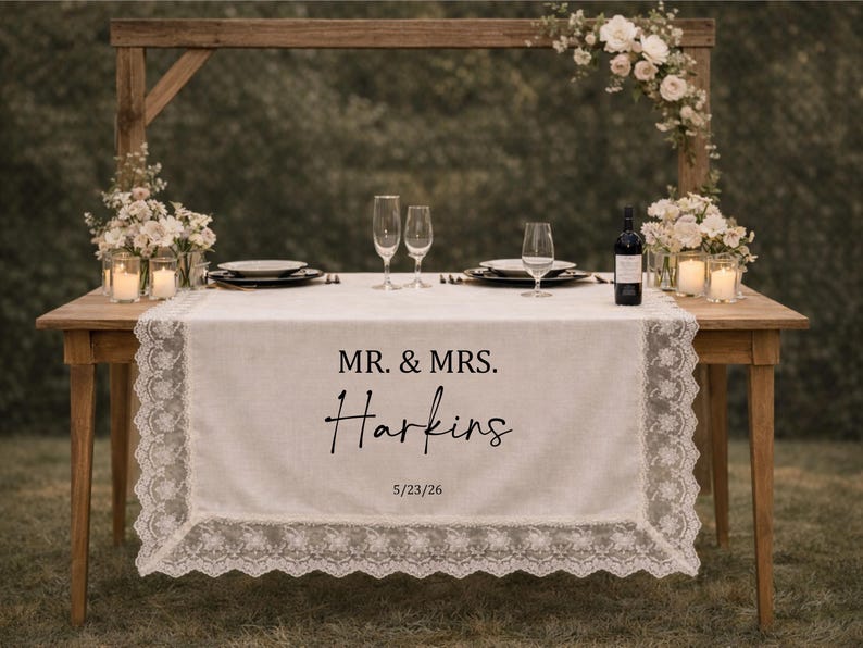 May include: A wedding reception table with a white lace-edged table runner bearing the text "MR. & MRS. Harkins" and the date "5/23/26". The table is set with champagne flutes, plates, and a wine bottle. Floral arrangements and candles decorate the wooden table.