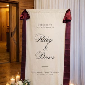 May include: A cream-colored wedding welcome sign with burgundy ribbon accents. The sign reads "WELCOME TO THE WEDDING OF Riley & Dean" with the date and location. Floral arrangements and candles flank the sign.