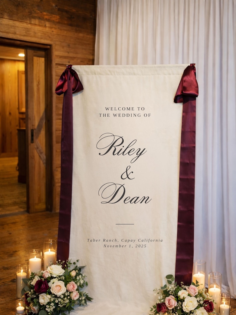 May include: A cream-colored wedding welcome sign with burgundy ribbon accents. The sign reads "WELCOME TO THE WEDDING OF Riley & Dean" with the date and location. Floral arrangements and candles flank the sign.