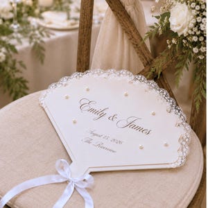 May include: A fan-shaped wedding programme made of off-white cardstock with a scalloped lace border and small pearl accents. The text reads Emily & James, August 15, 2026, and The Riverview. A white satin ribbon is tied at the base of the handle.