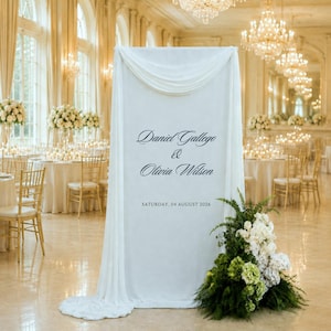 May include: A tall white fabric wedding sign featuring the black printed text Daniel Gallego & Olivia Wilson and the date SATURDAY, 04 AUGUST 2026. The fabric is draped at the top and adorned with a large arrangement of white flowers and green foliage.