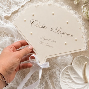 May include: A cream-coloured, fan-shaped card featuring white lace trim along the curved edge and small white pearls attached to the surface. The text reads Charlotte & Benjamin, August 15, 2026, and The Riverview. A white ribbon is tied at the base.