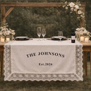 May include: A wooden table set for a special occasion, with a white lace-trimmed runner. The runner displays the text "THE JOHNSONS" and "Est. 2026". Champagne flutes, plates, and a wine bottle complete the elegant setup.
