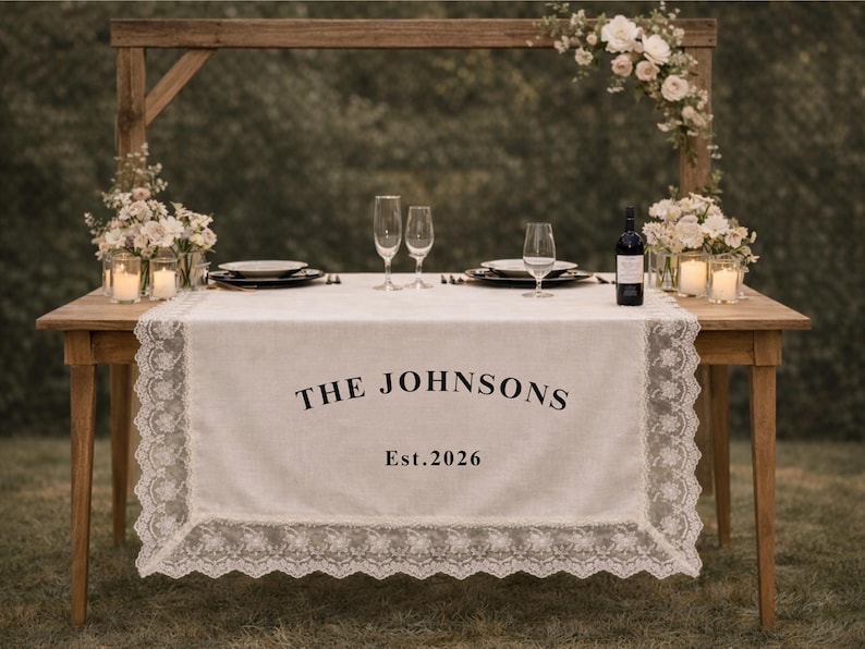 May include: A wooden table set for a special occasion, with a white lace-trimmed runner. The runner displays the text "THE JOHNSONS" and "Est. 2026". Champagne flutes, plates, and a wine bottle complete the elegant setup.