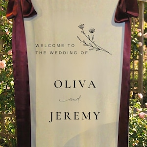 May include: A cream-colored wedding welcome sign with burgundy ribbons and the text "WELCOME TO THE WEDDING OF OLIVA and JEREMY" with floral accents. The sign also includes the location and date: Sicily, Italy, 6th September 2025.