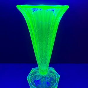 May include: A bright, glowing green glass vase with a flared top and a hexagonal base. The vase has a textured, etched design and stands on a blue surface. The vase is approximately 25 cm tall.