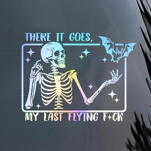May include: A holographic sticker featuring a skeleton with a bat and the text "THERE IT GOES, MY LAST FLYING F*CK". The skeleton is holding its hand out towards a bat. The sticker has a rainbow gradient.