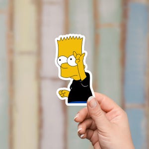May include: A die-cut sticker featuring Bart Simpson from The Simpsons cartoon. Bart is depicted with yellow skin, spiky yellow hair, a black shirt, and is making a hand gesture. The sticker is held by a hand against a blurred background.