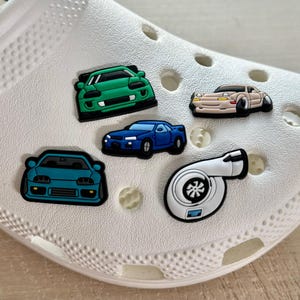 May include: A white shoe adorned with five colorful car-themed charms. The charms include a green sports car, a tan and white car, a blue car, a teal car, and a white turbocharger.