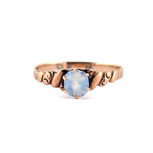 May include: A rose gold-toned ring featuring a central, round, pale blue cabochon stone. The band is crafted with a textured, twisted metal design that wraps around the finger, leading to the prong-set stone at the center of the ring's face.