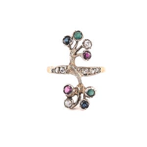 May include: A gold ring featuring a textured, branch-like design extending vertically from the band. The ring is set with multiple round-cut gemstones, including clear diamonds, deep blue sapphires, vibrant green emeralds, and pink rubies.