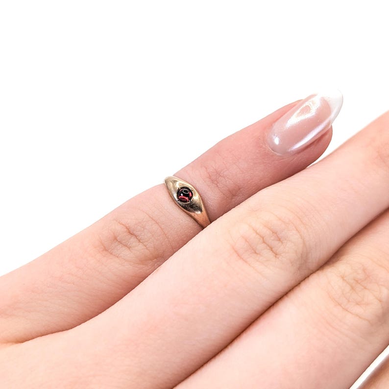 May include: A gold-toned ring featuring a smooth, tapered band and a single, small, round, dark red gemstone set securely in the center. The metal has a polished finish, and the design is simple, focusing on the contrast between the gold band and the stone.