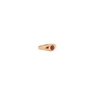 May include: A gold-toned metal ring featuring a smooth, polished band that tapers toward the center. The ring is set with a single, small, round, deep red gemstone held securely in a bezel setting at the top of the band's surface.
