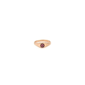 May include: A gold-toned metal ring featuring a smooth, polished band that tapers toward the center. The front of the ring is set with a single, small, round, deep red gemstone held securely in a simple, circular bezel setting.