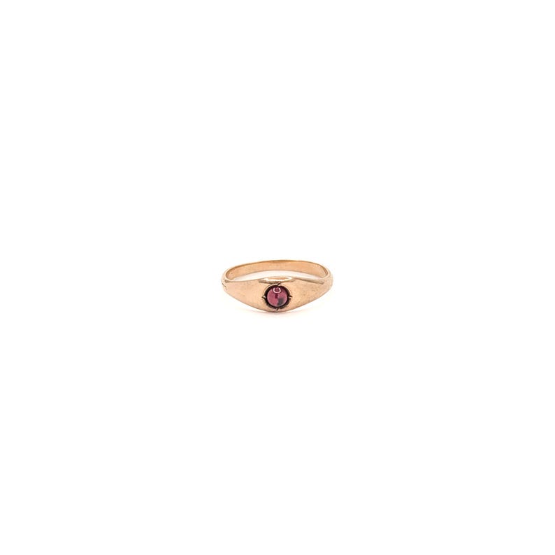 May include: A gold-toned metal ring featuring a smooth, polished band that tapers toward the center. The front of the ring is set with a single, small, round, deep red gemstone held securely in a simple, circular bezel setting.