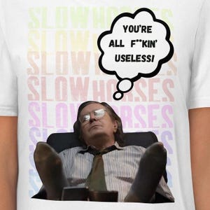 May include: White t-shirt with the text "SLOW HORSES" in a rainbow gradient pattern. The shirt features a man wearing glasses and a tie, with a speech bubble that says "YOU'RE ALL F**KIN' USELESS!"