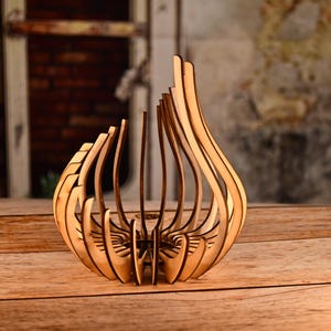 May include: A decorative wooden vase with a unique, open design. The vase is made of light brown wood and features a series of curved, vertical slats that create an organic, flowing shape. The vase is displayed on a wooden surface.