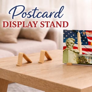May include: A postcard display stand with the text "Postcard DISPLAY STAND" in blue and red. The stand holds a postcard featuring the American flag, the Capitol Building, and an eagle. Two small wooden stands are in the foreground.