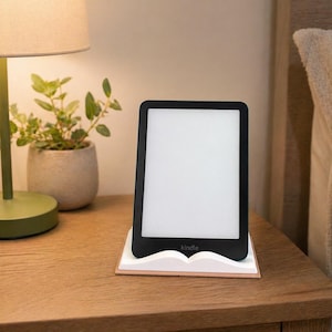 May include: A black Kindle e-reader with a white screen rests on a white and tan book-shaped stand. The e-reader is on a wooden nightstand next to a lamp and a potted plant.
