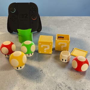 Könnte beinhalten: Eine Sammlung farbenfroher Gaming-Accessoires, darunter rote, gelbe und grüne Pilzfiguren, gelbe Fragezeichen-Boxen und ein schwarzer Controller. Die Artikel sind auf einer grauen Oberfläche vor blauem Hintergrund angeordnet.