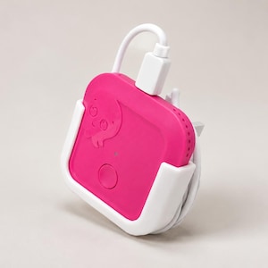 May include: A square, hot pink electronic device with a rounded top and a small circular button on the front. It features a moulded octopus graphic, is held within a white plastic cradle, and has a white charging cable plugged into the top.