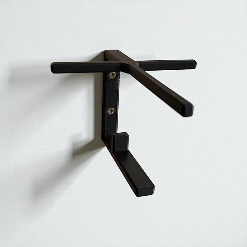 Puede incluir: Estante de pared de metal negro con un dise&ntilde;o &uacute;nico. El estante tiene un soporte vertical con dos brazos horizontales y un soporte inferior en &aacute;ngulo. Se fija a una pared blanca con tornillos.