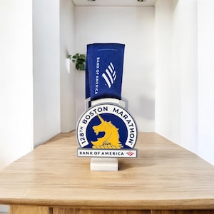 Peut inclure: Médaille du Marathon de Boston avec un ruban bleu et le logo Bank of America. La médaille présente une licorne jaune et le texte "128th Boston Marathon 2024". La médaille est présentée sur une surface en bois.