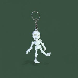 Puede incluir: Un llavero de esqueleto blanco con un llavero y una cadena plateados. El esqueleto está en una pose de caminar, con una pierna extendida. El fondo es verde oscuro liso.
