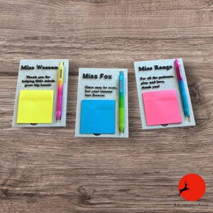 May include: Three personalized teacher gifts on a wooden surface. Each gift includes a notepad, a pen, and a message for "Miss Wesson", "Miss Fox", and "Miss Range". The pens have a gradient color scheme, and the notepads are yellow, blue, and pink.