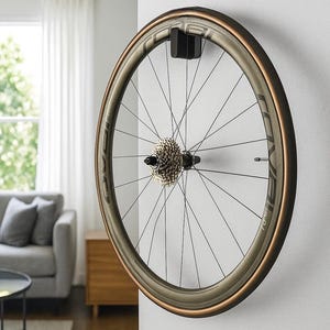 Peut inclure: Roue de vélo montée sur un mur blanc. La jante est de couleur bronze, avec une cassette argentée. Les rayons sont noirs, et le pneu a un flanc bronze. La roue est fixée avec un support noir.