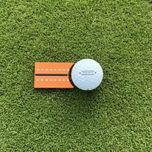 Peut inclure: Une balle de golf blanche à la surface alvéolée avec le texte TOUR SPEED imprimé en noir, placée à côté d'un outil d'alignement orange doté d'une ligne centrale noire flanquée de deux lignes blanches en pointillés sur sa surface rectangulaire.