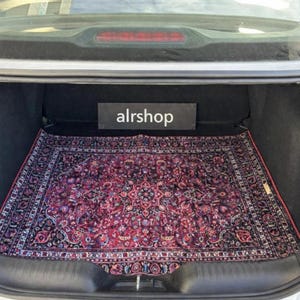 May include: A rectangular, patterned rug with a deep red and black color scheme, placed inside a car trunk. The rug features an intricate floral design. A sign above the rug reads "alrshop".