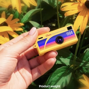Può includere: Una fotocamera giocattolo gialla con un design retrò, con una striscia arcobaleno e l'anno "1984". La fotocamera è tenuta in mano, con fiori gialli e foglie verdi sullo sfondo. La fotocamera ha un obiettivo nero e il testo "CAMERA Record Anytime Anywhere".