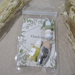 May include: A small clear glass bottle with a yellow liquid and a white label that says "Soap". The bottle is next to a silver and wooden diffuser with a white and green beaded charm. The charm says "Thank you".