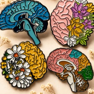 Könnte beinhalten: Vier Emaille-Anstecknadeln in Form menschlicher Gehirne. Eine ist blau mit dem Wort THINK, eine ist rosa mit violetten und gelben Blumen, eine ist gelb mit weißen Gänseblümchen und Bienen, und eine ist mehrfarbig mit rosa, orangen, grünen und braunen Abschnitten.