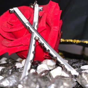 Könnte beinhalten: Nahaufnahme eines silbernen Metallobjekts in Form gekreuzter Schwerter, das auf einem Bett aus schwarzen und weißen Steinen ruht. Im Hintergrund eine leuchtend rote Rose, mit dunklem Hintergrund.