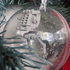 以下が含まれることがあります： 透明な球形のクリスマスオーナメントで、中に灰色のミニチュアAT-STウォーカーモデルが入っています。白いリボンで吊り下げられ、人工雪が詰められており、お祝いのホリデーデコレーションになっています。