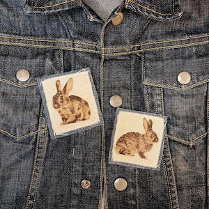 May include: A dark denim jacket with two square patches featuring brown rabbit illustrations. The patches are sewn onto the jacket's front, near the chest pockets. The jacket has silver buttons and yellow stitching.