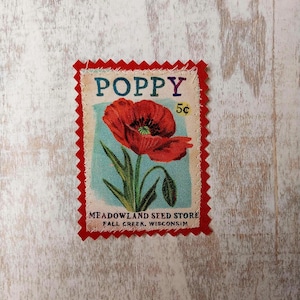 May include: A fabric patch featuring a red poppy flower with green leaves and the word "POPPY" in purple. The patch includes the text "MEADOWLAND SEED STORE FALL CREEK, WISCONSIN". The patch has a red, scalloped border.