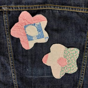 May include: Two fabric flower patches attached to a dark blue denim jacket. The top flower features pink gingham, blue floral, and white fabric segments. The bottom flower consists of white, pink gingham, and green floral patterned fabric pieces.