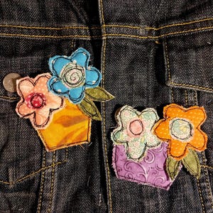 May include: Two fabric flower pot appliqués attached to a dark blue denim jacket. The left appliqué features a yellow pot with a pink and a blue flower. The right appliqué features a purple pot with a light green and an orange flower.