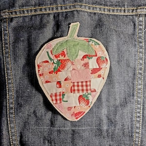May include: A strawberry-shaped fabric patch featuring a patchwork design of various pink, white, and red patterned squares, including strawberry prints and gingham checks. The patch has a green fabric leaf at the top and a light-colored border.