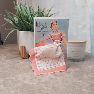 May include: A vintage-style greeting card featuring a woman in a pink dress, with a pink floral handkerchief attached. The card is labeled "Weigel's Pattern Book Summer 1/6". A white candle and a potted plant are in the background.