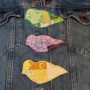 May include: A blue denim jacket featuring three bird-shaped fabric patches. The top patch is green with various patterns, the middle patch is purple with floral and gingham prints, and the bottom patch is yellow with orange and white floral designs.