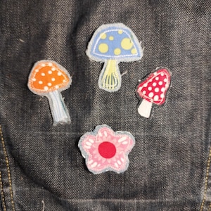 May include: Four fabric patches attached to dark denim fabric. The patches include an orange mushroom with white dots, a blue mushroom with yellow dots, a red mushroom with white dots, and a pink flower with a solid red center and white petals.