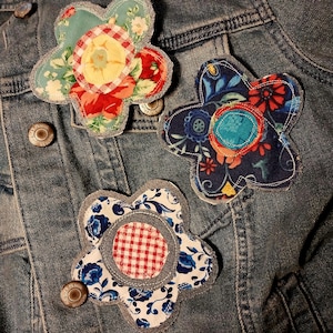 May include: Three handmade fabric flower brooches with denim edges rest on a denim jacket. The flowers feature various floral patterns in red, blue, and yellow, with a gingham center.