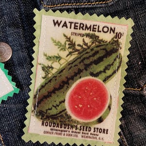 May include: A fabric patch with a watermelon design. The patch features a green and black striped watermelon and a red watermelon slice. Text on the patch reads "WATERMELON" and "ROUDABUSH'S SEED STORE". The patch is sewn onto a denim jacket.