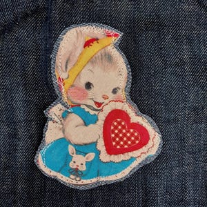 May include: A dark blue denim fabric featuring a stitched fabric patch of a cartoon rabbit wearing a yellow bonnet and a blue dress. The rabbit holds a large red heart with a white checkered center, and a small white rabbit is printed on the dress.